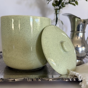 May include: A cylindrical ceramic canister with a light green speckled glaze and a matching removable lid with a central knob. The canister has a smooth, rounded base and is presented alongside a silver-toned metal pitcher and tray.