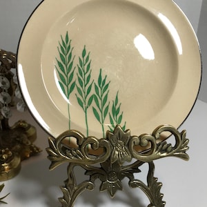 May include: A small, cream-colored ceramic plate with a green wheat design. The plate is on a gold metal stand.