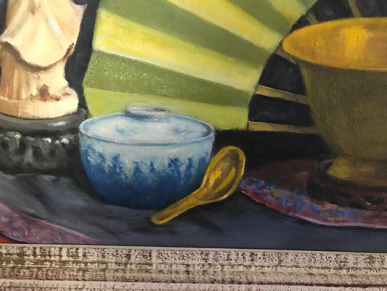 May include: A still life painting featuring a blue and white ceramic bowl, a gold spoon, a gold bowl, and a white statue on a black base. The background is a yellow and green fan.
