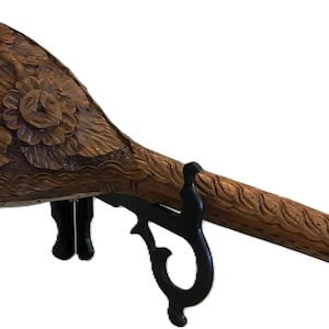 May include: A hand-carved wooden club with floral carvings on the head and a textured handle. The club is displayed on a black metal stand. The wood has a warm, brown tone, and the carvings are detailed.