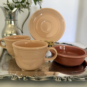 May include: A collection of ceramic tableware including two light peach-colored cups, one matching saucer, and one terracotta-colored bowl. These items are arranged on a silver-toned metal tray alongside a silver-toned metal pitcher.