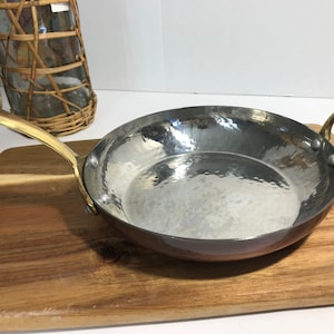May include: A hammered copper and stainless steel pan with brass handles. The pan is on a wooden cutting board.
