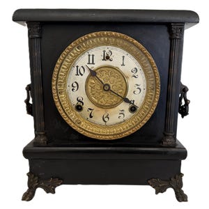 Puede incluir: Un reloj de chimenea antiguo negro con una esfera redonda. La esfera tiene números romanos negros y manecillas doradas. El reloj es rectangular con columnas decorativas y patas ornamentadas.