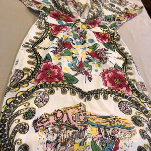 以下が含まれることがあります： 鮮やかな花柄とペイズリー柄の白いドレス。ドレスは大きなピンクの花、緑の葉、装飾的なボーダーが特徴です。デザインには、人物と馬がいるメリーゴーランドのシーンが含まれています。ドレスは半袖です。