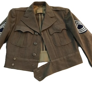 Puede incluir: Chaqueta militar verde oliva con cuello con solapas, cuatro bolsillos delanteros y cierres de botones. La chaqueta presenta parches en las mangas, incluyendo galones y un emblema circular. Prenda de estilo vintage.