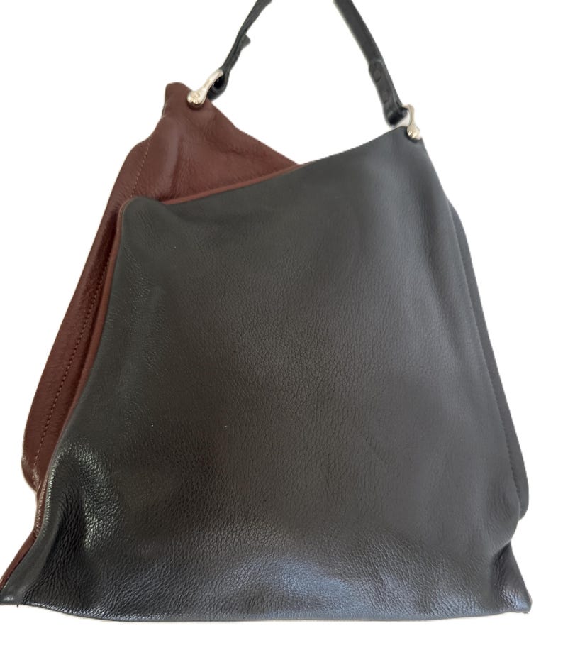 Puede incluir: Un bolso de hombro de cuero de dos tonos. El exterior es marr&oacute;n oscuro, mientras que el interior es marr&oacute;n claro. El bolso tiene una sola correa negra y herrajes plateados.
