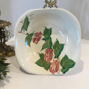 May include: A white ceramic bowl with a square shape and rounded edges. The bowl features a hand-painted design of pink flowers and green leaves.