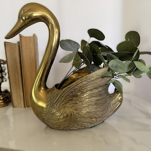 Peut inclure: Un cache-pot en laiton poli en forme de cygne avec des gravures détaillées de plumes sur le corps. Le cygne possède un long cou courbé et un centre creux contenant actuellement plusieurs tiges de feuilles d'eucalyptus vertes et arrondies.