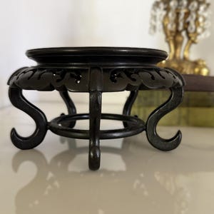 May include: A dark brown, circular wooden display stand featuring a smooth, flat top surface and an intricate, openwork carved apron. The stand is supported by four curved legs that terminate in scrolled feet, connected by a central ring.