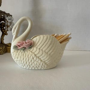 May include: Cream-colored ceramic swan match holder with a textured wing design and a pink rose accent. The swan's open back holds a bundle of wooden matches. A decorative item for the home.