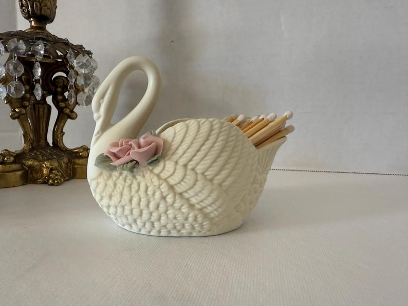 May include: Cream-colored ceramic swan match holder with a textured wing design and a pink rose accent. The swan's open back holds a bundle of wooden matches. A decorative item for the home.