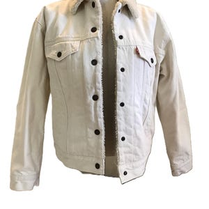 May include: Cream-colored denim jacket with a light-colored sherpa collar. The jacket has dark brown buttons and two chest pockets. A small red tag is visible on the left chest pocket. The jacket is displayed on a mannequin.