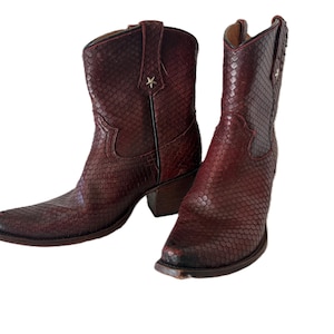 May include: A pair of burgundy-colored cowboy boots with a snakeskin pattern. Each boot has a pointed toe, a small star accent, and a stacked heel. The boots are made of a textured material.