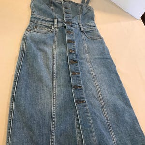 Peut inclure: Une robe en jean bleu clair avec un devant boutonn&eacute; et des bretelles fines. La robe a un corsage ajust&eacute; et une jupe l&eacute;g&egrave;rement &eacute;vas&eacute;e.