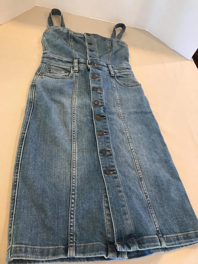 Peut inclure: Une robe en jean bleu clair avec un devant boutonn&eacute; et des bretelles fines. La robe a un corsage ajust&eacute; et une jupe l&eacute;g&egrave;rement &eacute;vas&eacute;e.