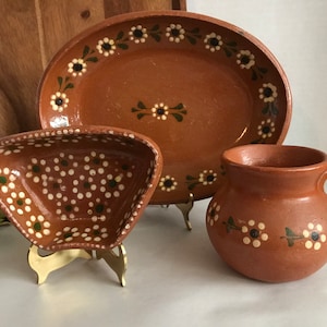 May include: Set of three terracotta pottery dishes with white and green floral designs. The dishes are on a wooden surface.