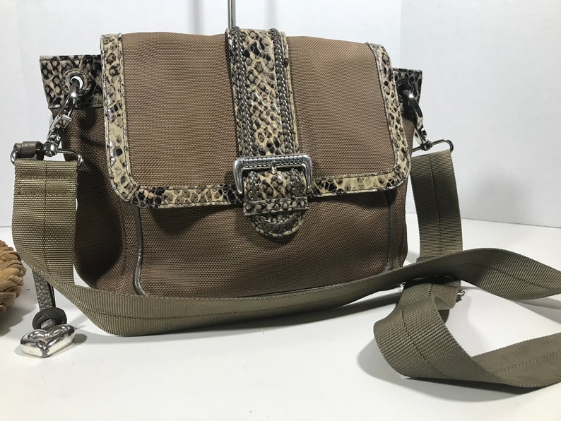 Puede incluir: Un bolso bandolera de lona marr&oacute;n con una solapa con estampado de piel de serpiente y una hebilla plateada. El bolso tiene una correa larga ajustable y un peque&ntilde;o amuleto unido a la correa.