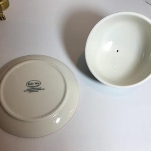 May include: A white ceramic bowl and a matching plate with the text "Dutch Wax" on the bottom. The bowl is round with a small black dot in the center. The plate is also round.