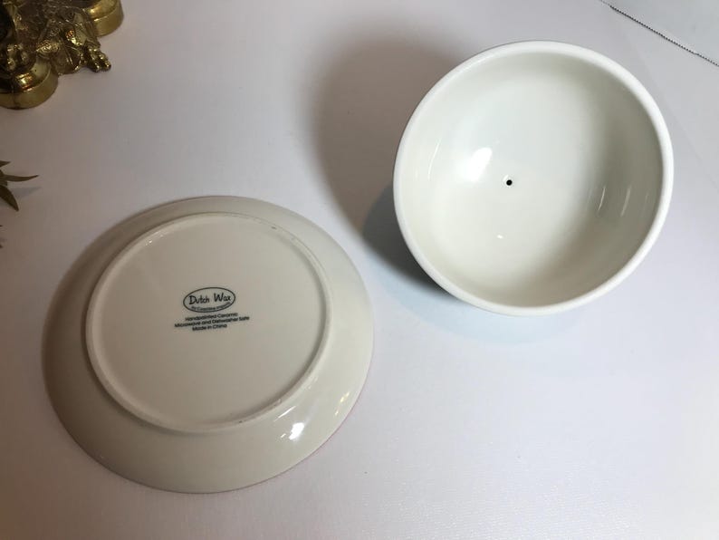 May include: A white ceramic bowl and a matching plate with the text "Dutch Wax" on the bottom. The bowl is round with a small black dot in the center. The plate is also round.
