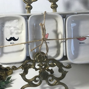 Puede incluir: Un juego de tres platos de cerámica blanca con un diseño decorativo. Los platos están atados con un cordel y tienen las palabras "His", "Ours" y "Hers" escritas en ellos. Los platos están sobre un soporte de metal dorado.