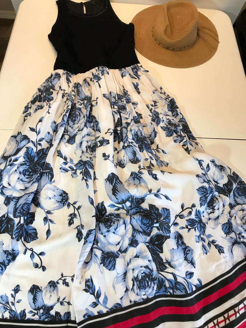 May include: A black sleeveless dress with a white skirt patterned with blue flowers. A straw sun hat sits beside the dress. The skirt has a black and red striped border.