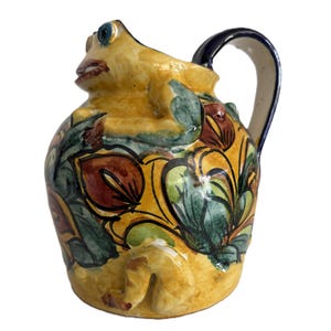 May include: A ceramic pitcher in the shape of a frog, with a yellow body and a dark blue handle. The pitcher is decorated with green, red, and yellow floral patterns.