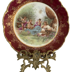 May include: A decorative plate with a claret rim and gold floral detailing, depicting a pastoral scene with cherubs and figures. The plate is presented on an ornate gold-coloured metal stand, exhibiting a vintage style.