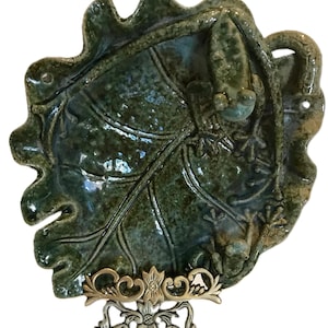 May include: A ceramic leaf-shaped dish with a glossy green glaze, featuring embossed leaf veins and small frog figurines. The dish is mounted on a decorative metal stand with floral and scrollwork details. The overall design is nature-inspired.