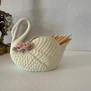 May include: Cream-colored swan-shaped match holder with a pink rose accent. The ceramic holder is filled with wooden matches. The swan has detailed wing and feather patterns. A decorative gold candle holder is in the background.