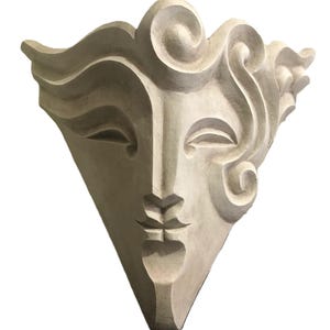Könnte beinhalten: Eine cremefarbene, dreieckige Wandskulptur mit einem stilisierten Gesicht mit geschlossenen Augen, einer kleinen Nase und einem geschlossenen Mund. Die Skulptur hat fließende, wirbelnde haarähnliche Details an der Oberseite und den Seiten.