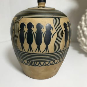 May include: A tan ceramic jar with a lid, featuring black hand-painted designs of stylised figures holding hands in a circle. The vessel is decorated with horizontal black lines at the top and bottom, and a scalloped pattern near the base.