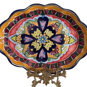 May include: A colorful, hand-painted ceramic serving platter with a scalloped edge. The oval dish features a central floral design in blue, yellow, and purple, surrounded by red, orange, and green patterns. The platter is displayed on a decorative gold-colored metal stand.