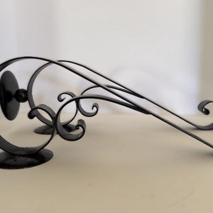 May include: Two black metal wall-mounted brackets featuring a curved, scroll-like design. Each bracket has a circular base plate for mounting and a decorative ball finial at the end of the scroll, constructed from smooth, dark-finished metal.