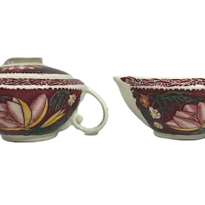 Puede incluir: Un azucarero vintage con tapa y una jarrita para la crema. Ambas piezas están decoradas con un diseño floral en tonos burdeos, rosa, amarillo y verde. Los bordes tienen un borde decorativo. El azucarero tiene dos asas.