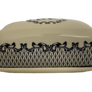 Puede incluir: Mantequera de cerámica color crema con tapa. La fuente y la tapa están decoradas con un patrón geométrico azul marino y adornos ornamentados. La fuente se asienta sobre una base rectangular a juego.