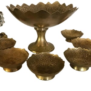 Puede incluir: Un juego de cuencos de servicio de latón antiguo y un cuenco con pedestal. Los cuencos tienen un patrón floral decorativo y bordes festoneados. El cuenco con pedestal está sostenido por una base redonda.