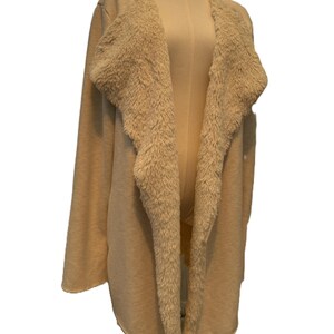 Può includere: Cardigan beige aperto sul davanti con colletto in morbida ecopelliccia. Il maglione a maniche lunghe è esposto su un manichino. Il cardigan è di colore neutro, adatto per la stratificazione e l'abbigliamento casual.