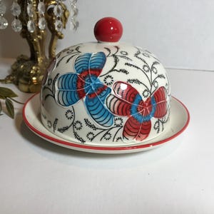 May include: A white ceramic butter dish with a domed lid and a matching plate. The lid is decorated with red and blue floral designs and a red knob. The plate and lid have a red trim.