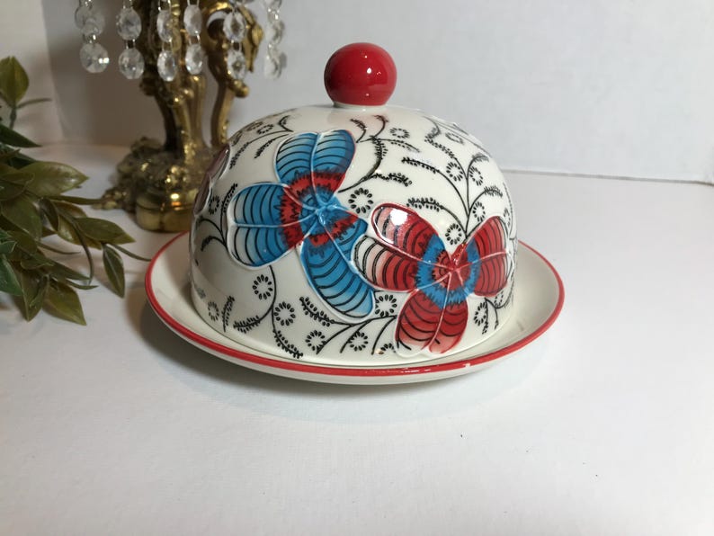 May include: A white ceramic butter dish with a domed lid and a matching plate. The lid is decorated with red and blue floral designs and a red knob. The plate and lid have a red trim.