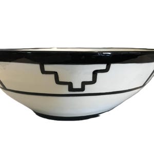 May include: A white ceramic bowl with a black geometric design. The bowl has a wide, shallow shape with a black rim and a black stepped pattern around the middle. The design is symmetrical and appears to be hand-painted.