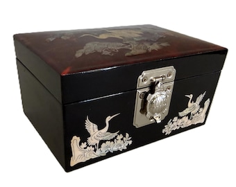 May include: A rectangular black lacquered box featuring white inlaid crane and floral motifs on the front and a dark reddish-brown lid with similar inlay. The box includes a silver-toned metal latch shaped like a turtle on the front panel.