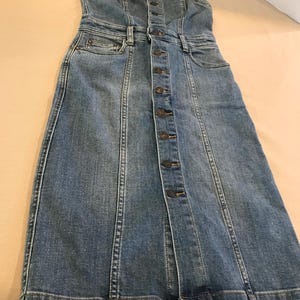 Peut inclure: Une robe en jean bleu avec un devant boutonn&eacute; et des bretelles fines. La robe a un corsage ajust&eacute; et une jupe l&eacute;g&egrave;rement &eacute;vas&eacute;e.
