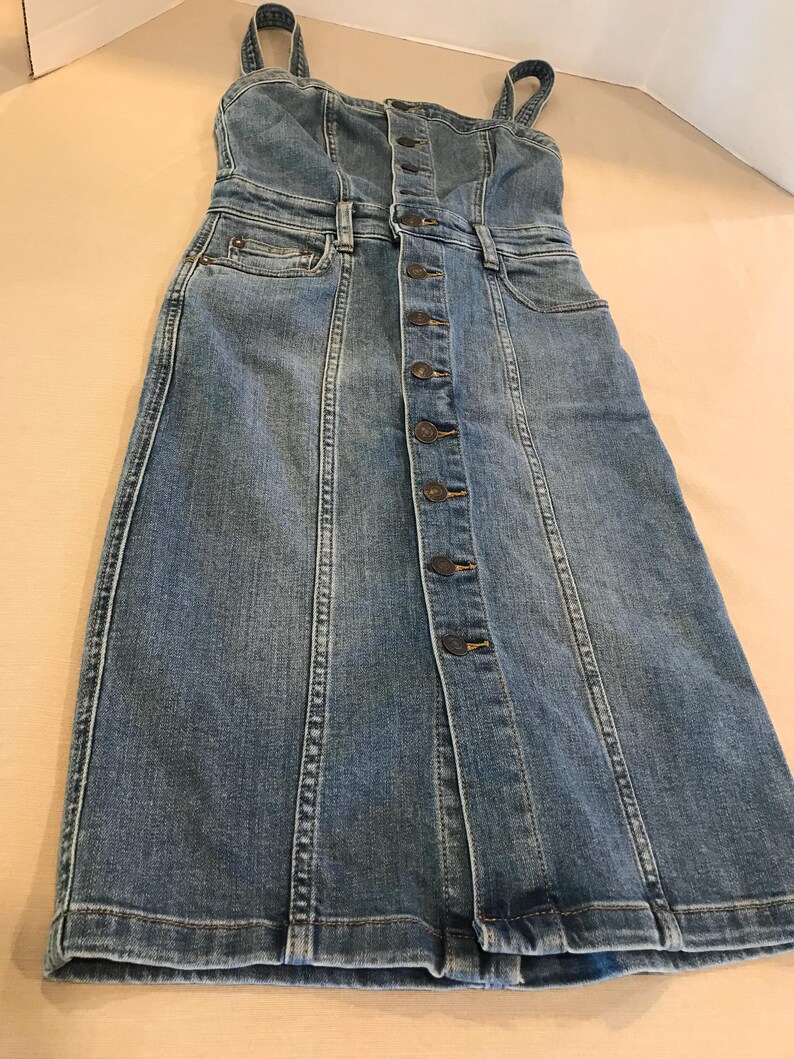 Peut inclure: Une robe en jean bleu avec un devant boutonn&eacute; et des bretelles fines. La robe a un corsage ajust&eacute; et une jupe l&eacute;g&egrave;rement &eacute;vas&eacute;e.