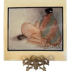 May include: A framed watercolour print depicting a seated figure wrapped in a blanket with red, green, and brown tones. The artwork is presented on a decorative gold-coloured metal stand, highlighting the artistic details and presentation.