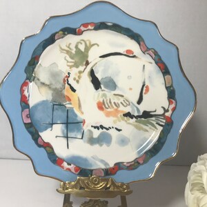 May include: A blue and white ceramic plate with a watercolour design of a bird and flowers. The plate has a scalloped edge and a gold rim.