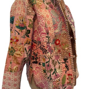 May include: A quilted jacket in shades of pink, featuring a floral patchwork design. The jacket has a mandarin collar, long sleeves, and two front pockets. The edges are trimmed with a contrasting striped fabric. The jacket is a lightweight garment.