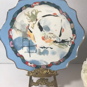 May include: A blue and white ceramic plate featuring a watercolour design of a bird and flowers. The plate is displayed on a gold metal easel.