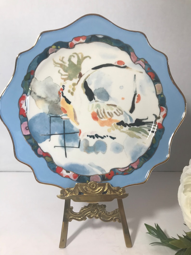 May include: A blue and white ceramic plate featuring a watercolour design of a bird and flowers. The plate is displayed on a gold metal easel.