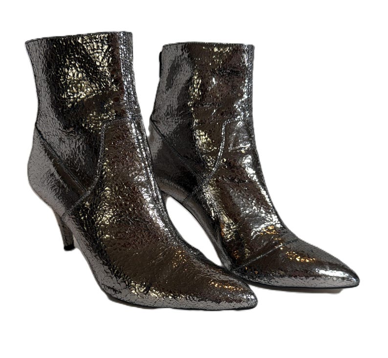 May include: A pair of metallic silver ankle boots with pointed toes and stiletto heels. The boots have a textured, crinkled appearance and a zipper on the back. The heels measure approximately 7.6 cm.