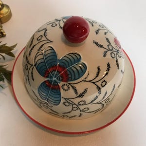 May include: A white ceramic butter dish with a domed lid and a matching plate. The lid features a large blue and red floral design, a red knob, and black vine-like accents. The plate has a red rim.